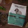 A bag of Science Selective Adult Rabbit Food is placed on a wooden surface, surrounded by natural ingredients such as green peas, hay, grains, and pellets. The packaging features a white and green design with an image of a lop-eared rabbit. Small bowls and wooden scoops hold various ingredients, emphasizing the food's natural composition.
