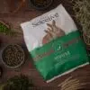 A bag of Science Selective House Rabbit Food is placed on a wooden surface, surrounded by natural ingredients such as green peas, hay, linseeds, and pellets. The packaging features a white and green design with an image of two rabbits. Small bowls and wooden scoops hold various ingredients, emphasizing the food's natural composition.
