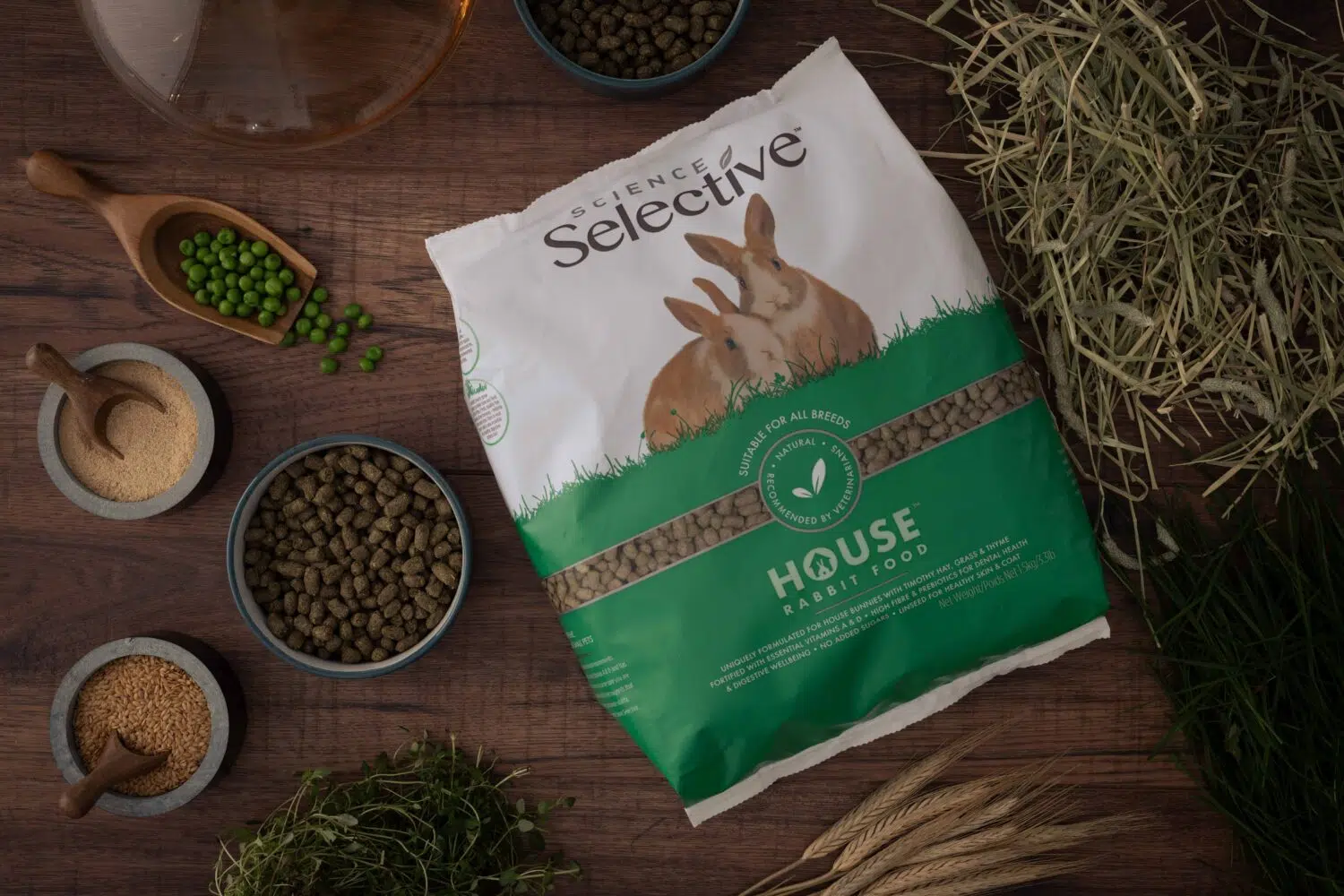 A bag of Science Selective House Rabbit Food is placed on a wooden surface, surrounded by natural ingredients such as green peas, hay, linseeds, and pellets. The packaging features a white and green design with an image of two rabbits. Small bowls and wooden scoops hold various ingredients, emphasizing the food's natural composition.