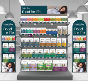 A retail gondola end display for Science Selective pet foods. The display features shelves filled with bags of small pet food, neatly arranged by color-coded packaging for different species and life stages. The top header panel reads 'Science Selective – Food for life.' and includes a photo of a woman smiling while cuddling a grey rabbit. Both side panels also display the 'Science Selective – Food for life.' branding, along with the line 'For every stage in life.' and images of various product packs for rabbits, guinea pigs, hamsters, and other small pets. The overall design uses teal, white, and accent colors from the product range, creating a clean and expert look under soft retail lighting.