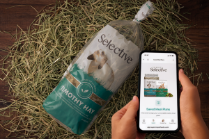 An overhead shot shows a person holding a smartphone that displays the "Saved Meal Plans" page of the Science Selective website. The phone is held over a wooden surface covered in loose hay, next to a large clear bag of Science Selective Timothy Hay featuring a picture of a lop-eared rabbit.