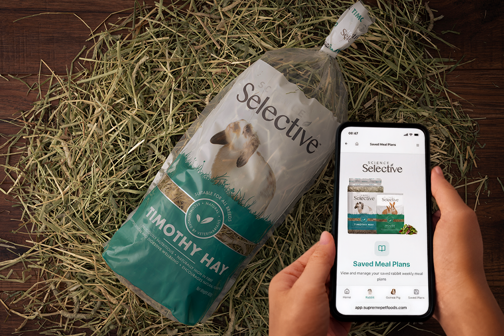 An overhead shot shows a person holding a smartphone that displays the "Saved Meal Plans" page of the Science Selective website. The phone is held over a wooden surface covered in loose hay, next to a large clear bag of Science Selective Timothy Hay featuring a picture of a lop-eared rabbit.