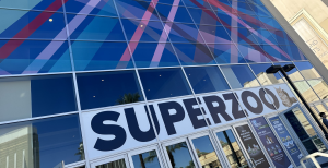 A large glass-paned entrance to the SuperZoo pet industry trade show featuring a bold "SUPERZOO" logo banner with a cat graphic over the doors. The upper windows are covered in a vibrant, overlapping abstract pattern of blue, pink, and red diagonal stripes, with reflections of palm trees and nearby buildings visible in the lower glass panels.